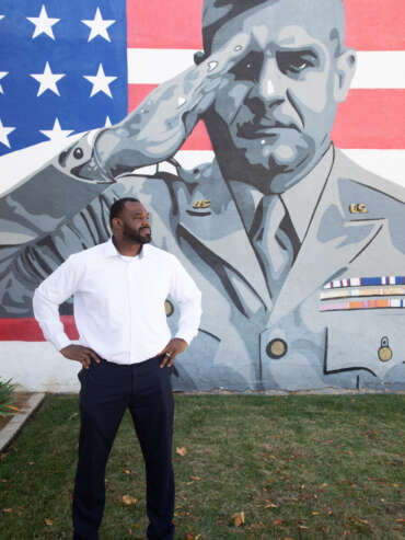 Getro Elize posing in front of a mural of a US veteran saluting against an American flag