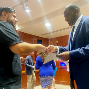 Getro Elize in a suit hands paperwork to another man in a meeting room.